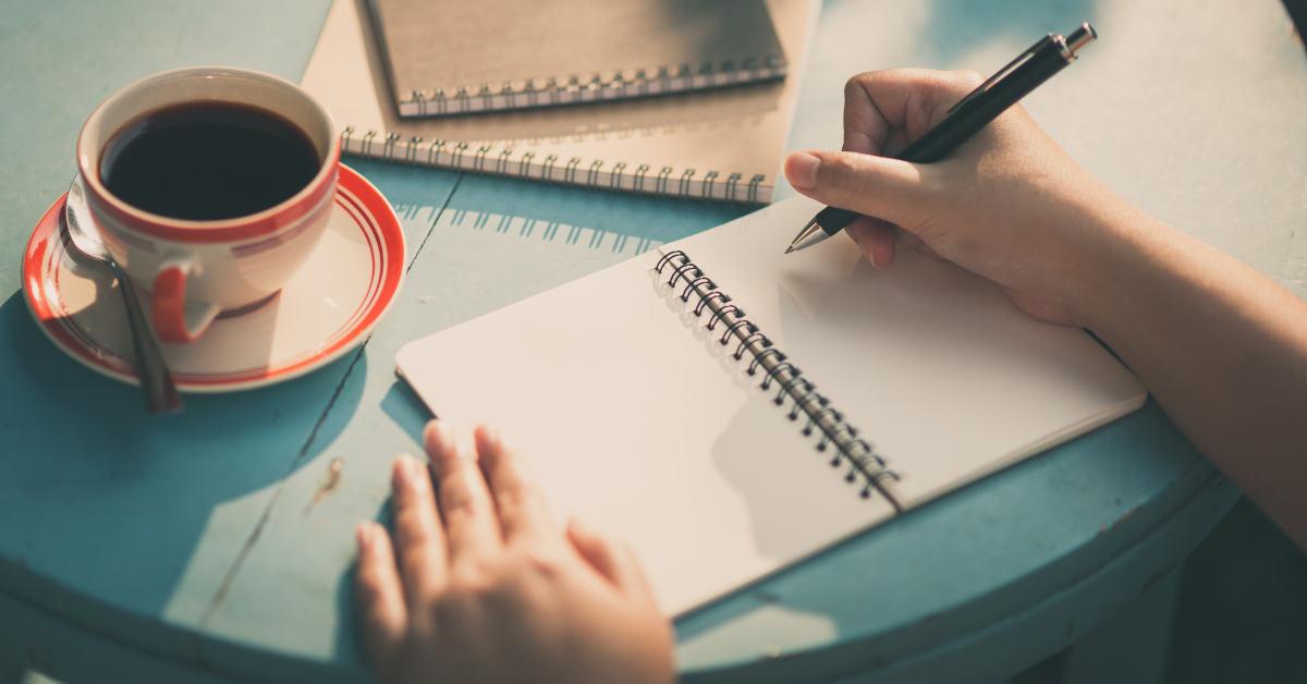 Donna alla scrivania che scrive su un quaderno a spirale e una tazza di caffè a fianco