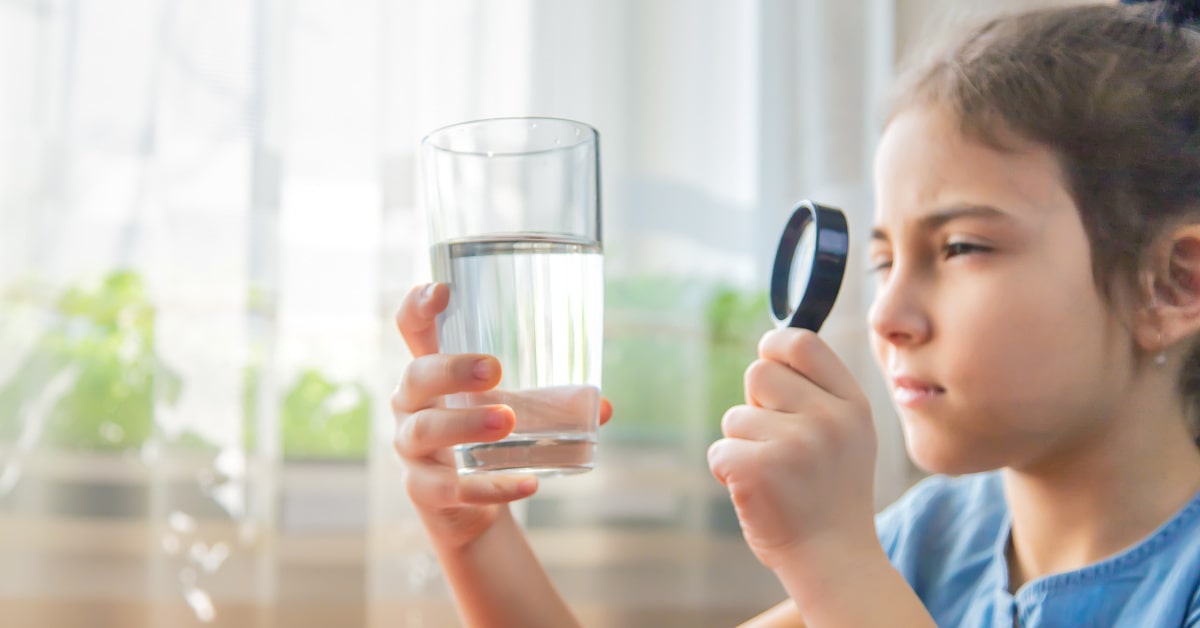 Bambina con un bicchiere di acqua in mano mentre osserva con una lente di ingrandimento