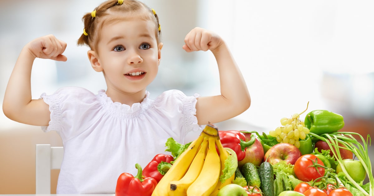 Bambina che fa il segno di forza con le braccia con frutta e verdura sul tavolo