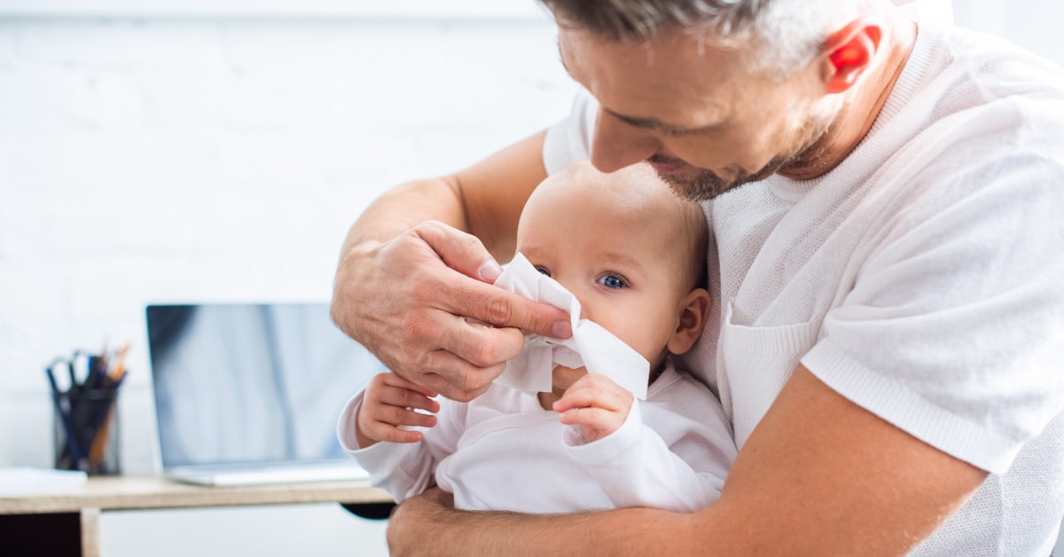 Babbo con figlia neonata in braccio mentre le pulisce il naso con un fazzoletto di carta