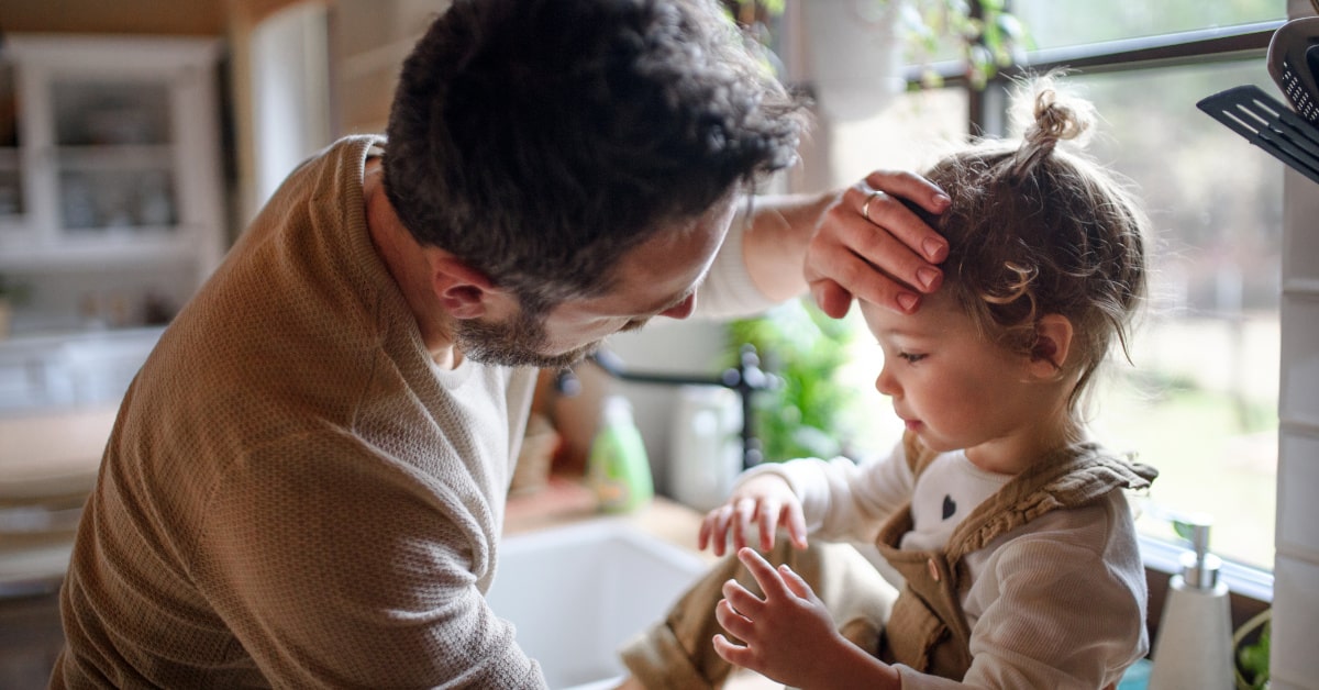 Bambina seduta sul lavello di casa, mentre il papà le sente la fronte con la mano per misurare la temperatura