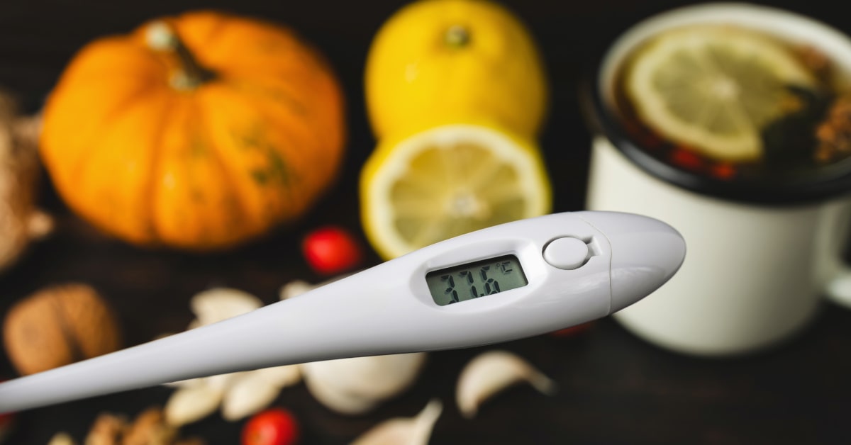 Termometro digitale che indica 37.6 °C e sullo sfondo rimedi naturali e tisana calda