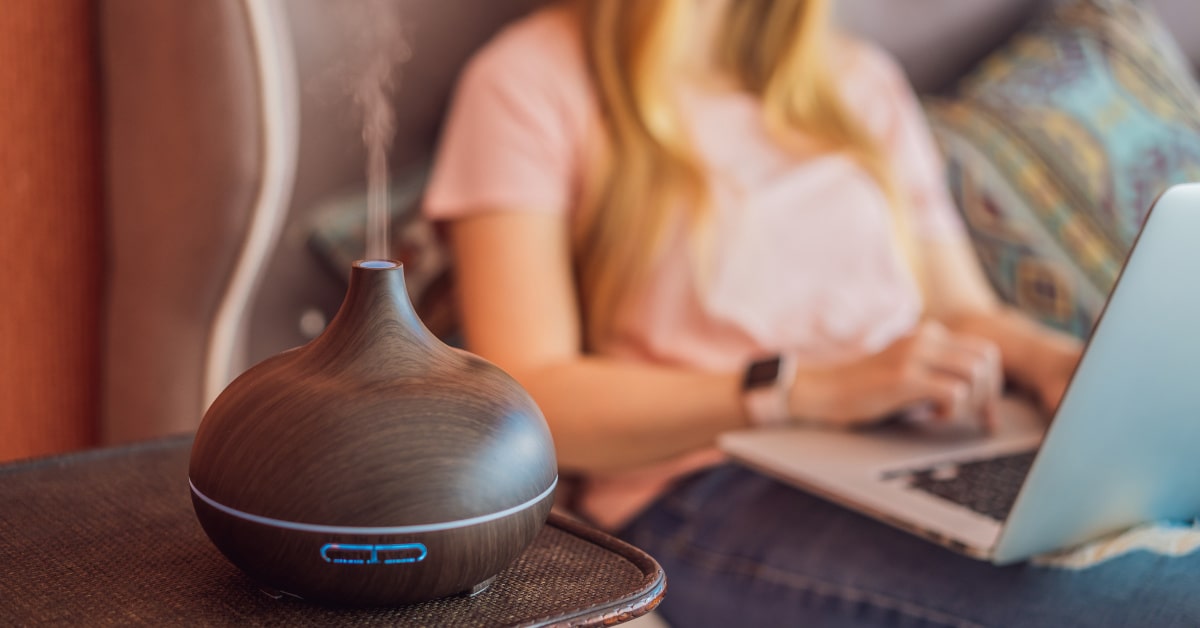 Diffusore di oli essenziali posizionato in salotto con donna sullo sfondo che lavora al PC